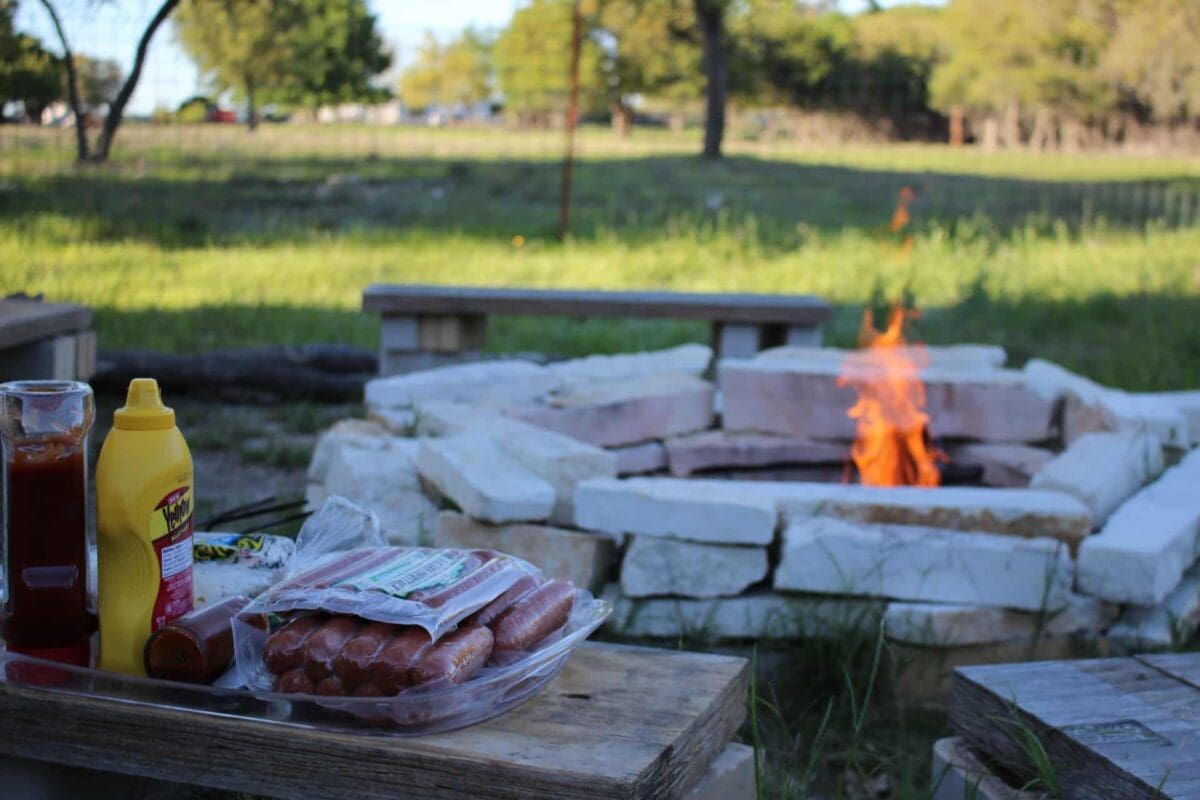 An outdoor fire pit made from stacked stone. A fire is burning in the pit, and a nearby table has hot dogs, drinks, and condiments.